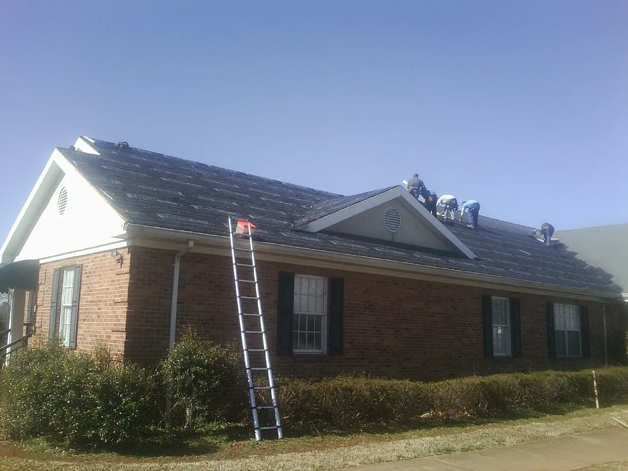Crew tearing shingles off a brick ranch roof with a ladder set — Metal Roof Repair work by our crew in Lone Jack