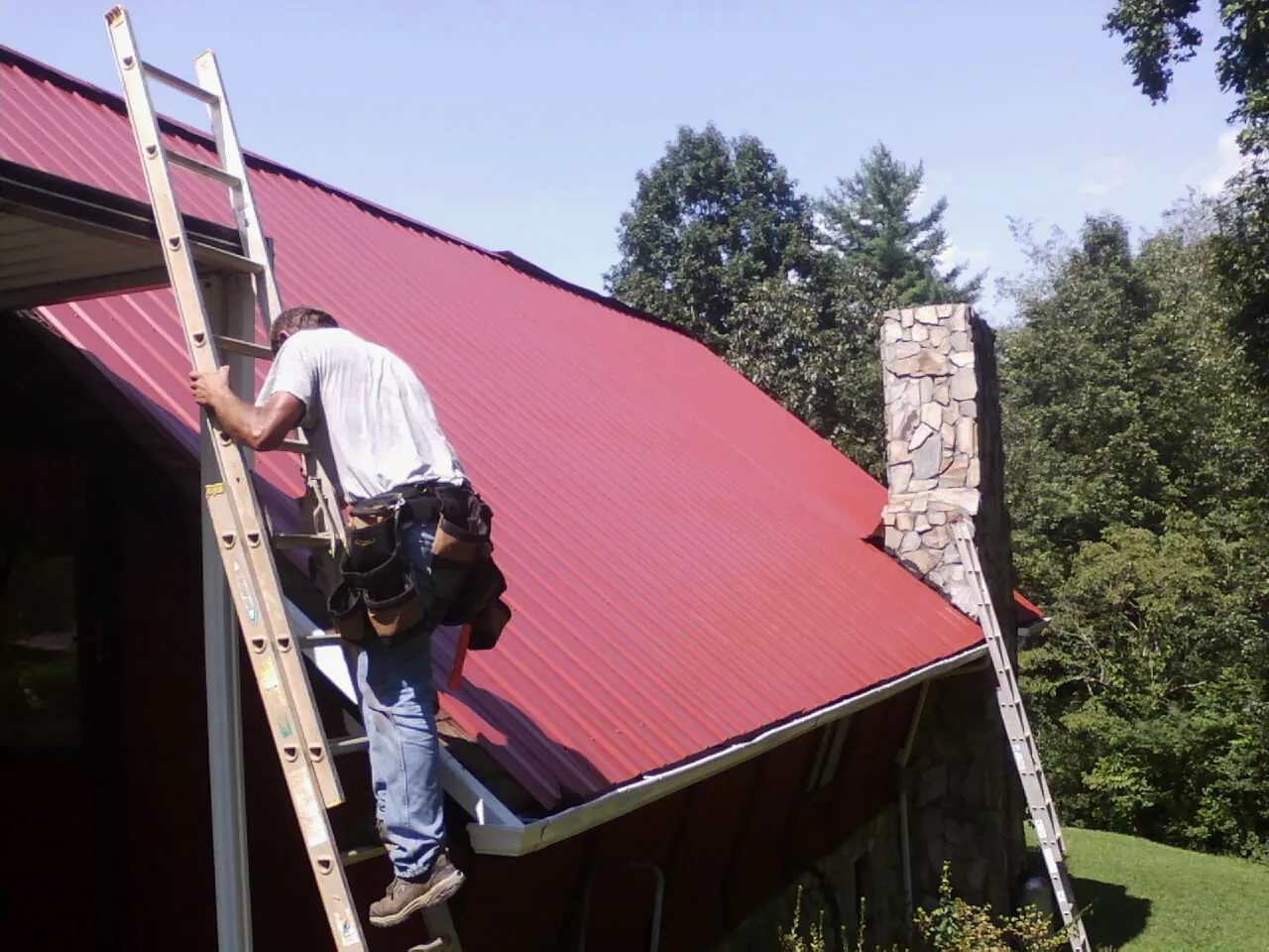 Roofer on a ladder beside a red metal roof and stone chimney — Soffit & Fascia Repair by Roofing Brothers in Lone Jack